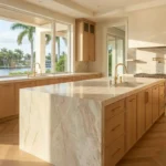 Kitchen Spacious waterfront kitchen in Boca Raton featuring natural wood cabinetry, waterfall marble island, brass fixtures, and large windows overlooking the water.